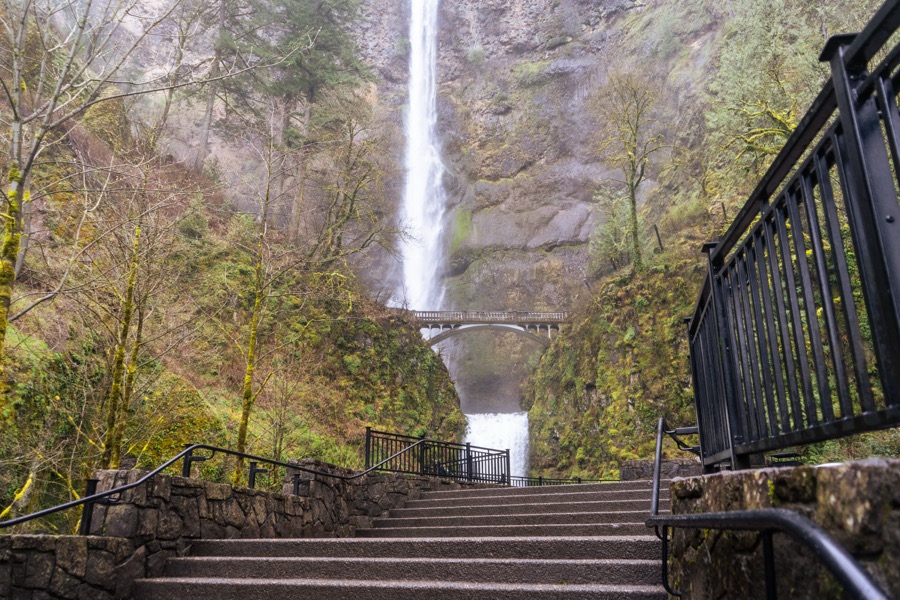 A waterfall and footbridge viewed from stone steps in a wooded gorge.