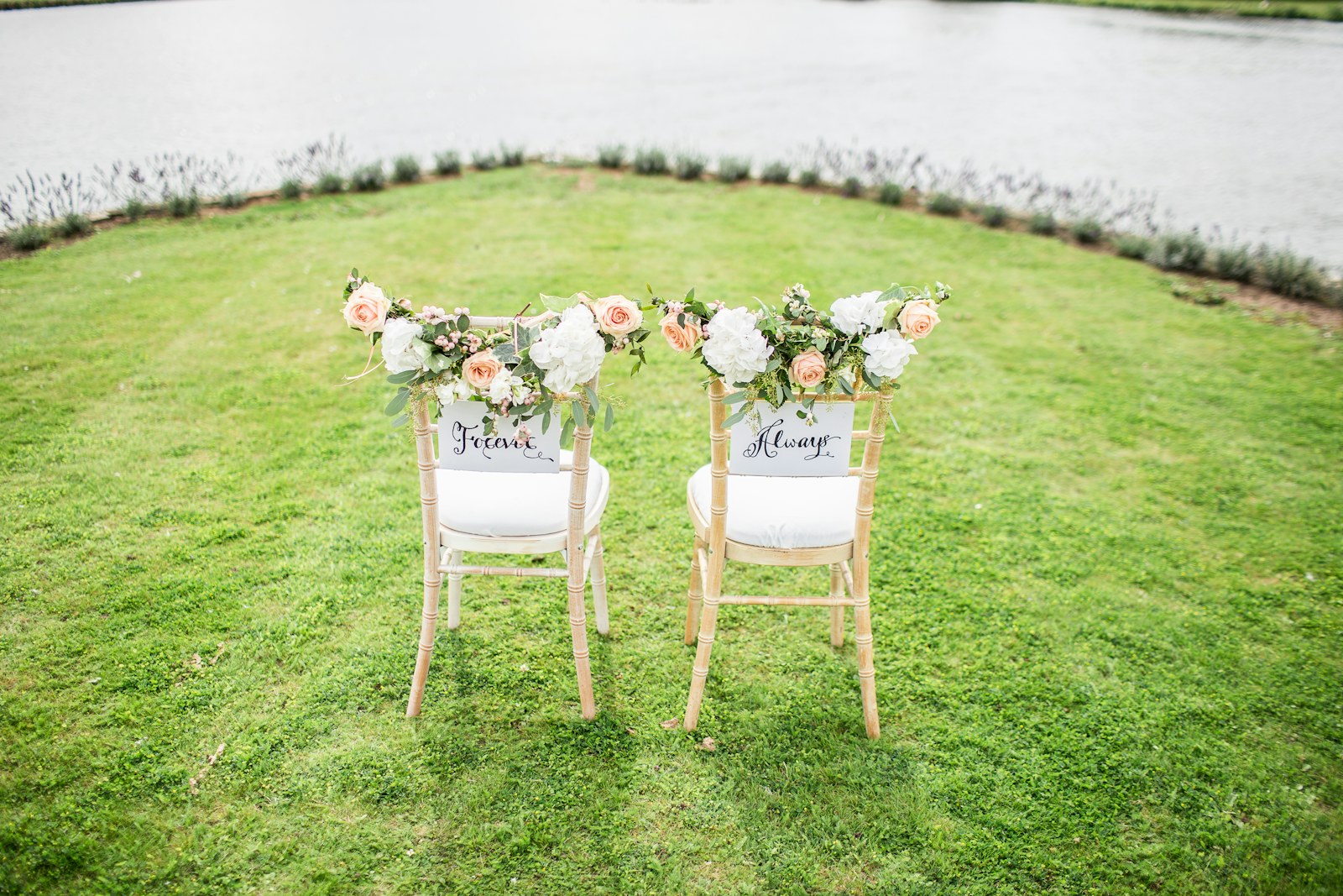 Two flower-decorated chairs on a lawn beside the water.